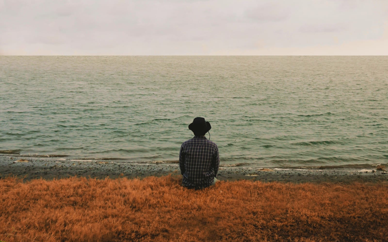 A man in a plaid shirt and bucket hat sits alone on a grassy shore, facing a vast grey-green lake under a cloudy sky.